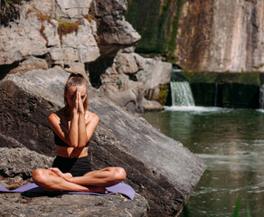 A beautiful tanned girl with a sports figure is doing yoga exercises against the backdrop of a picturesque waterfall in the summer. Sits in the lotus position. She is wearing black shorts and a top.