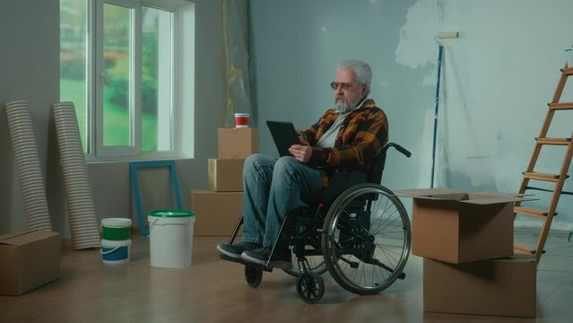 An elderly disabled man moves in a wheelchair plans renovation using a digital tablet. A pensioner checks building materials. Room with window, ladder, cardboard boxes, wallpaper rolls, paint bucket.