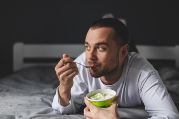 Handsome man lies on his bed while eating a pint of pistachio ice cream with spoon. He is look at window, dreaming. Eating in bed. Happy beautiful man resting in his comfortable bed at home. Yummy.