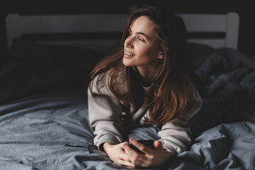 Fototapeta premium Young happy smile woman day dreaming think looking up hold cell phone on bed in bedroom, at home, girl lying type message, look at window, flirting.