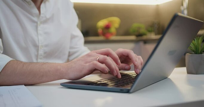 Close up male hands of unrecognizable male man at table typing computer keyboard online. Man typing on a laptop while sitting in the kitchen. Internet technologies concept.