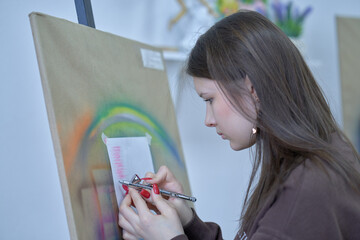 A teenage girl, an aspiring artist, paints with an airbrush on paper. Drawing lesson in an art studio.
