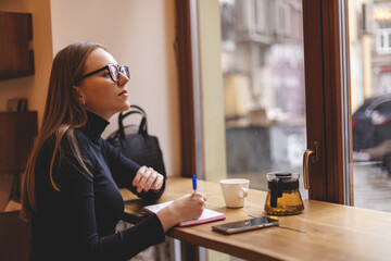 Pondering attractive young woman with pen in hand thinking on plans and writing list to do in...