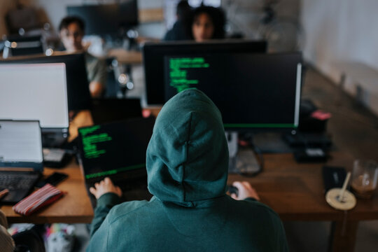 Rear view of young male hacker wearing hood coding on computer at desk in creative workplace