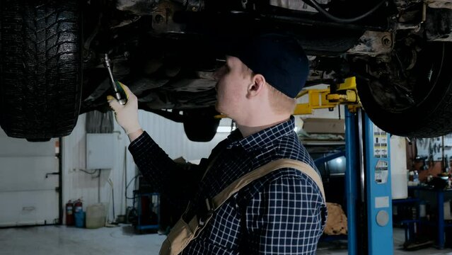 A young mechanic in uniform works in a car service with a lifted car. Car repair and maintenance