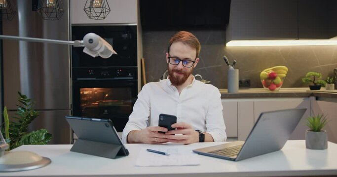 Attractive businessman, handsome successful freelancer typing text into smartphone and reading news on screen, sitting at table on the home kitchen background.