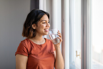 Smiling Young Arab Female Drinking Water While Standing Near Window At Home