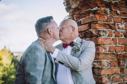 Bald gay man kissing groom on mouth by brick wall at wedding ceremony