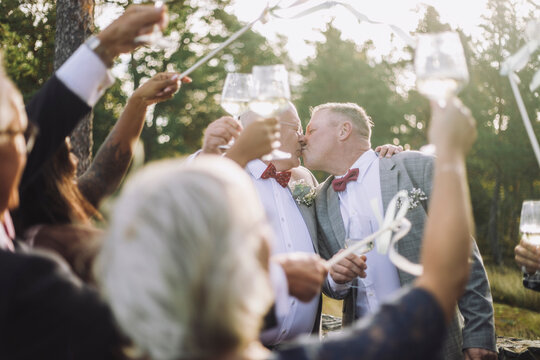 Newlywed gay couple kissing in front of guests raising toasts at wedding ceremony