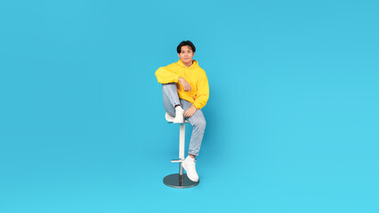 Fototapeta na wymiar Korean Teenager Boy Sitting On Bar Stool Over Blue Background
