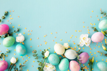 Spring Easter background with pastel colorful easter eggs, flowers and green eucalyptus decorated golden confetti on blue table. Top view photo.