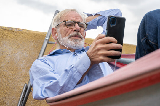 Mature business man with gray hair and beard in blue shirt and jeans laying in hammock with smartphone on terrace
