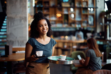 Happy black waitress serving coffee in cafe and looking at camera.