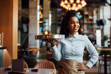Happy black waitress with tray in cafe looking away.