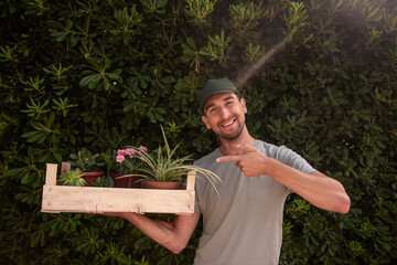 Man gardener in green cap holds wooden box with houseplants in front of living evergreen fence Phillyrea latifolia. Delivery of seedlings from plant nursery. Small business, hobby, gardening. Mock up