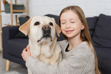 happy girl with closed eyes embracing labrador dog at home.