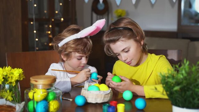 Easter Family traditions. Two caucasian happy school children kids with bunny ears paint and decorate eggs with paints for holidays while sitting together at home table. Kids having fun together