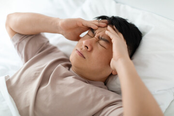 Fototapeta na wymiar Unhappy asian man lying in bed, touching head, closeup