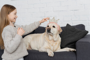happy preteen girl adjusting toy crown on labrador lying on couch.