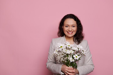 Attractive dark haired middle aged multiethnic woman, wearing stylish elegant gray jacket, holding cute bouquet of chrysanthemums and gypsophila flowers, smiling at camera, isolated on pink background
