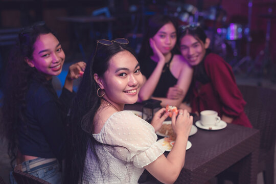 Four lovely Southeast Asian ladies dining in a tavern with live band. Emphasis on a young beautiful woman dressed in a white puffed top beaming at the camera while eating seafood dishes.