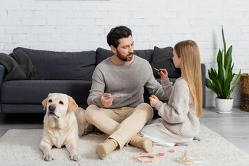 girl holding lip gloss while sitting near father and labrador on carpet.
