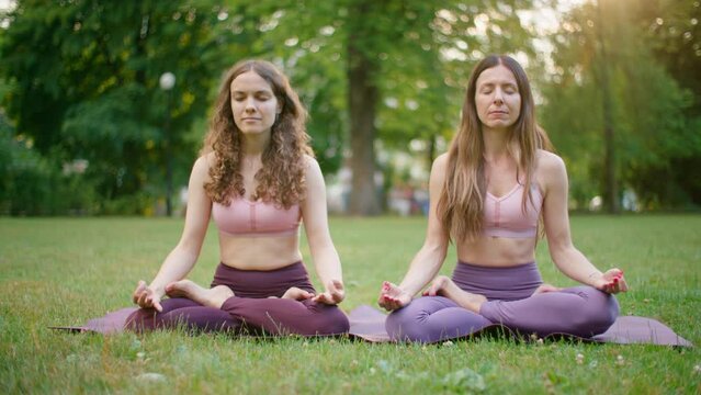 Two Sporty Women doing Meditaton in Park. Fit Ladies Stretch and do Sports outdoors in Lotus Poses. Mindfullness and Healthy Lifestyle and Meditation 4K wide orbit shot