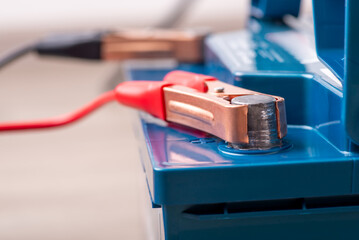 Red and black clamps connected to positive and negative terminals of blue car lead-acid battery, close up. Transmission of electricity, charging process, care of automobile