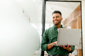 Confident male entrepreneur is standing in a modern office, typing away on his laptop. His cheerful...