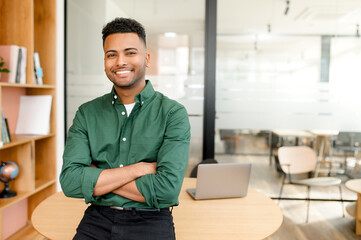 Ambitious and proficient Indian male employee in casual attire leans on the office desk, smiling...