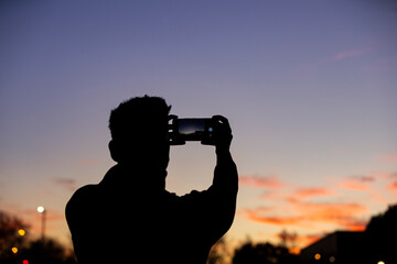 Rear silhouette of man taking a photo with smart phone at sunset in the city