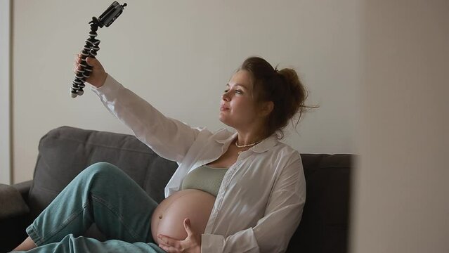 Happy expecting mother and popular vlogger is recording video for online blog sits on sofa in modern flat and showing her big naked belly looks at smartphone camera. Pregnancy and blogging concept.