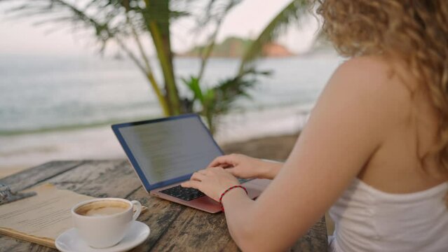 Young female developer working on laptop by the ocean. Woman freelancer coding at outdoor tropical cafe. Caucasian girl working remotely typing on computer at exotic location. Close-up backview shot