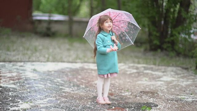 A beautiful little girl with brown hair and a toothless smile, wearing a coat under the umbrella, looking up on the falling white petals. Spring footage. Soft focus. 