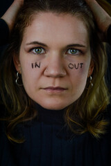 Fototapeta premium making decisions - portrait of a beautiful blonde woman with the words in and out written on her cheeks and looking intently in uncertainty