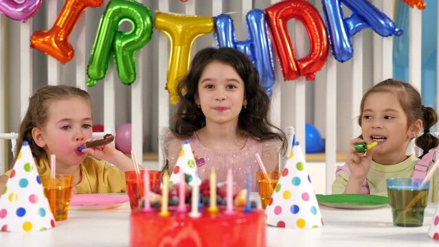 Cheerful children blow pipes at birthday parties while sitting at a table with a delicious red cake with candles. Birthday party with balloons and tinsel.