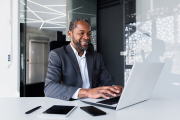 Successful african american manager inside office at workplace, man working using laptop smiling and happy with achievement results and financial money, boss in business suit typing on keyboard.
