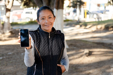 Hispanic woman holding a cell phone in the park - Mayan woman showing her phone screen - cell phone mockup - obrazy, fototapety, plakaty