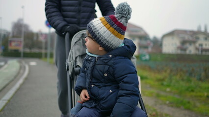 Parent pushing child seated on stroll in city sidewalk. Kid wearing winter clothes. Child wears warm clothes beanie and coat strolling outdoors