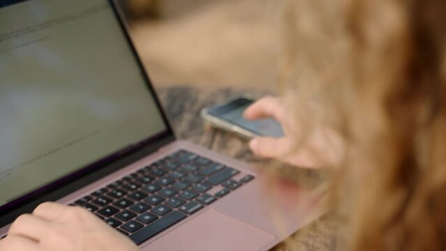 Female developer typing on laptop takes phone with diagrams and checks statistics. Freelancer woman working remotely on computer, typing and checking colorful charts on smartphone. Remote work concept