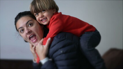 Obraz premium Little boy jumping on mother back. Authentic real life parent and child relationship laughing and smiling. Mom and kid embrace