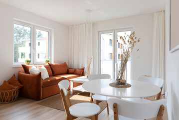 Modern interior of living room and dining room in open space. Round table with chairs and stylish red sofa with pillows in cozy room with window and wooden floor. New apartment in hotel.
