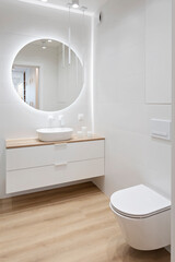 Luxury bathroom with round mirror with led lights, stylish washbasin and wooden floor. Modern interior of bathroom with wc and bath. Vertical.