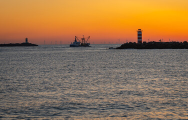 Fishing boat in silhouette shortly after sunset in between two lighthouses of south and north pier Ijmuiden, The Netherlands