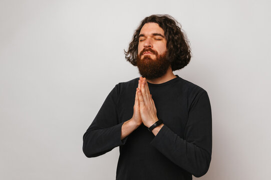 Young bearded christian man with long hair holds hands together for a prayer.