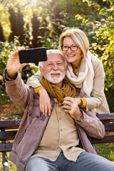 Happy senior couple relaxing together in a city park, sitting on a park bench and using a smartphone to take a selfie of themselves