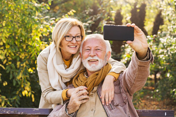 Happy senior couple relaxing together in a city park, sitting on a park bench and using a smartphone to take a selfie of themselves