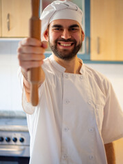 Portrait of a young bearded chef smiling while holding a rolling pin looking at camera