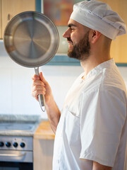 Side portrait of a young bearded chef smiling while showing a stainless pan