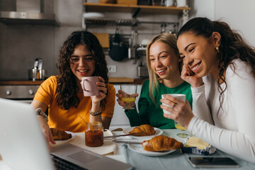 Three female friends are sitting in the kitchen and looking at laptop
